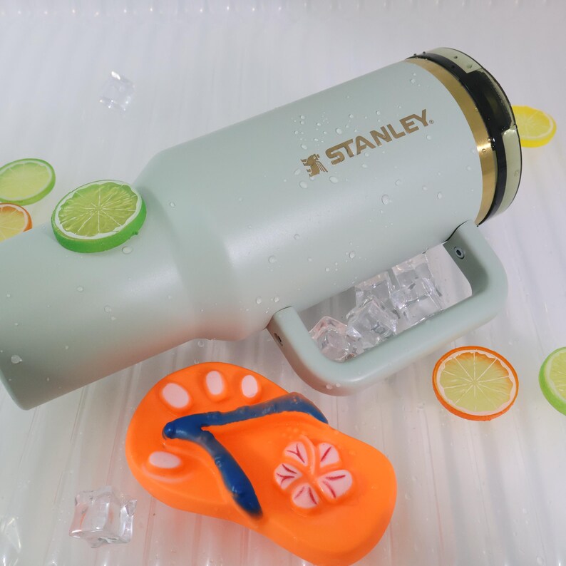 Op de afbeelding: Een lichtgroene Stanley waterfles met een handvat, ijsblokjes en het Stanley-logo. De fles is versierd met citroen- en limoenschijfjes en een oranje teenslipper.