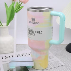 May include: A Stanley Quencher H2.0 tumbler with a pastel tie-dye design in pink, yellow, and blue. The tumbler has a light blue handle and a white label with the brand name and product details. The tumbler is next to a white vase with pink flowers.