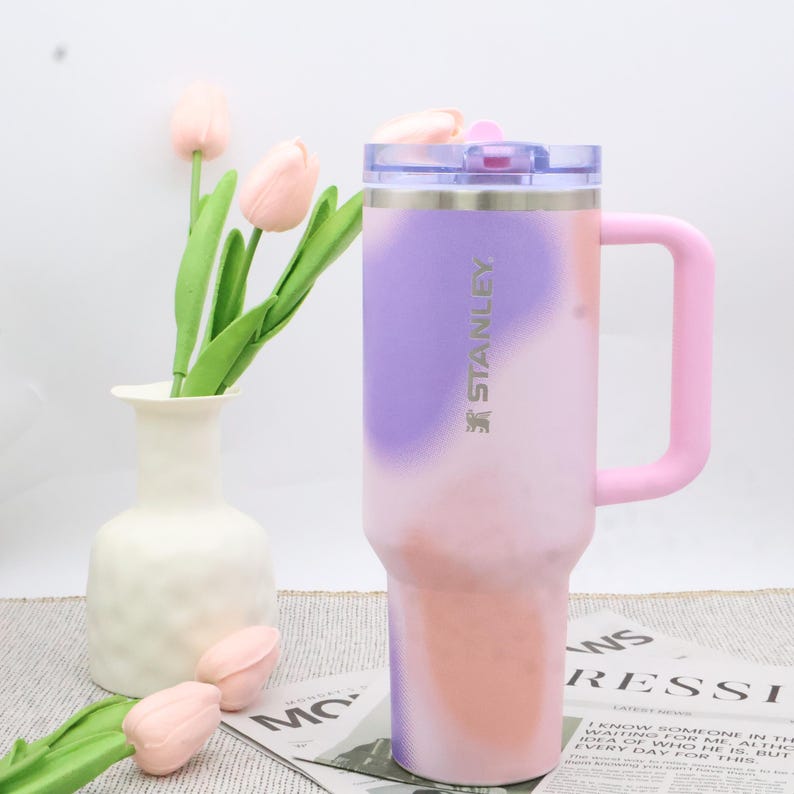 Puede incluir: Un vaso Stanley de acero inoxidable con un dise&ntilde;o degradado en tonos morado, rosa y melocot&oacute;n. Tiene un asa rosa y una tapa transparente. El logotipo de Stanley es visible. Un jarr&oacute;n blanco con tulipanes rosas est&aacute; en el fondo.