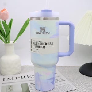 May include: A Stanley Quencher H2.0 tumbler with a pastel tie-dye design in shades of blue, purple, and white. It has a handle, a lid, and a straw. The tumbler is labeled with the brand name and product details.