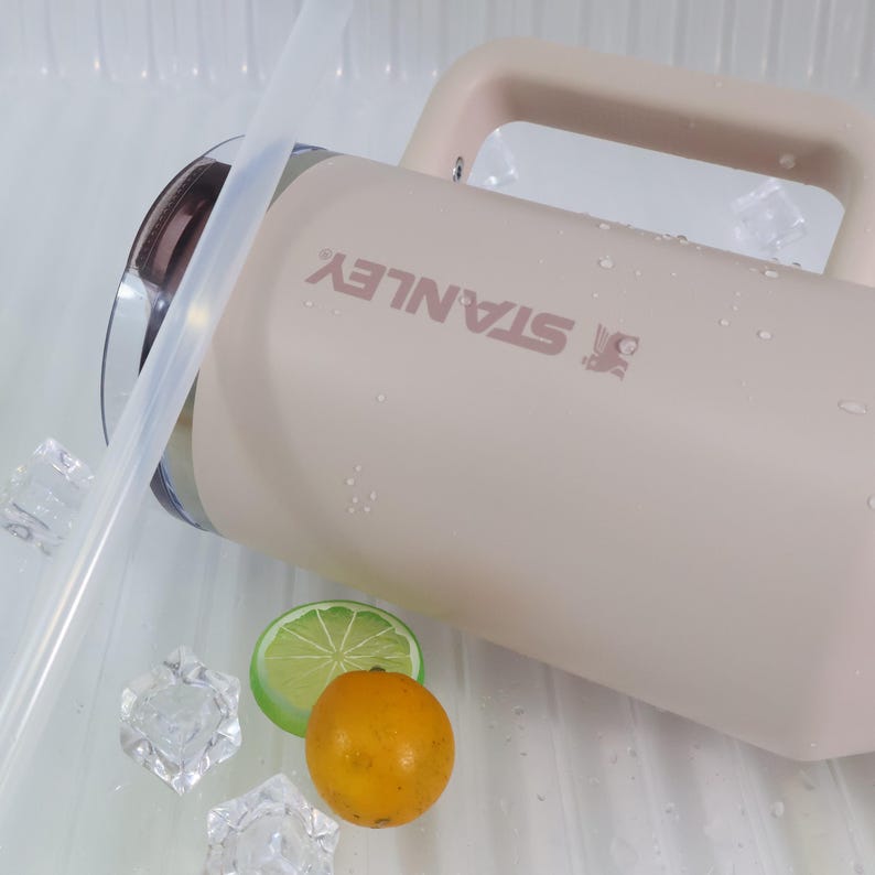 Peut inclure: Une gourde Stanley beige avec une poign&eacute;e et le nom de la marque imprim&eacute; en rose. Une paille transparente, des gla&ccedil;ons, une tranche de citron vert et une petite orange sont &agrave; c&ocirc;t&eacute; de la gourde. La gourde est pos&eacute;e sur une surface blanche.