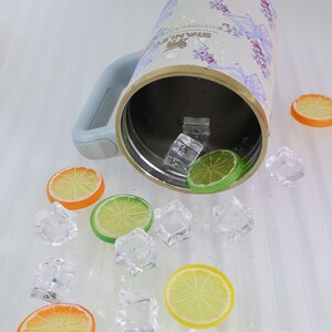 May include: A stainless steel insulated tumbler with a floral design, a light blue handle, and a gold rim. The tumbler is surrounded by ice cubes and slices of orange and lime.