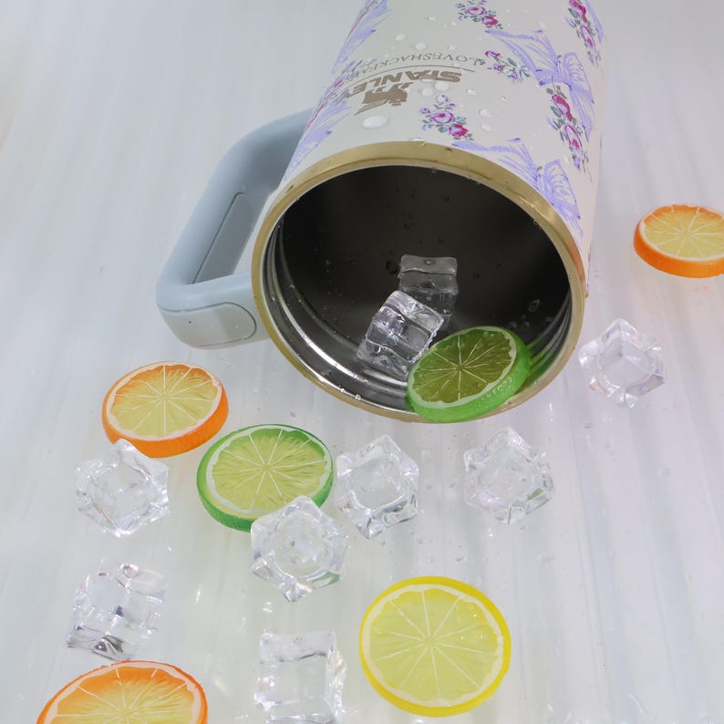 May include: A stainless steel insulated tumbler with a floral design, a light blue handle, and a gold rim. The tumbler is surrounded by ice cubes and slices of orange and lime.