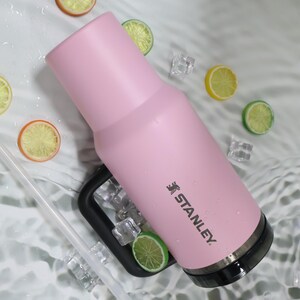May include: A pink Stanley tumbler with a black handle and base, featuring the Stanley logo. The tumbler is surrounded by ice cubes and citrus fruit slices, creating a refreshing aesthetic. A clear straw is also visible.