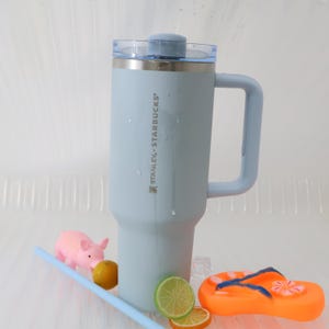 Puede incluir: Un termo de viaje Stanley Starbucks azul claro con asa y tapa transparente. La taza est&aacute; junto a un cerdo rosa, una pajita azul, un lim&oacute;n, rodajas de lima y una chancla naranja.