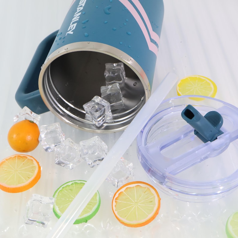 Puede incluir: Un vaso t&eacute;rmico de color verde azulado con asa, cubitos de hielo y una pajita transparente. El vaso tiene rayas rosas y la palabra "STANLEY" impresa. La tapa es transparente con una boquilla verde azulado. Rodajas decorativas de lim&oacute;n y naranja esparcidas.