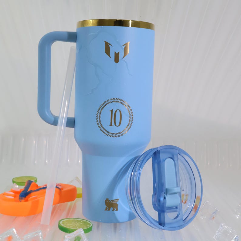May include: A light blue insulated tumbler with a gold rim, handle, and logo. The tumbler has a clear lid and straw. The number "10" is printed on the tumbler. A small orange toy and lime slices are in the foreground.