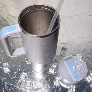 May include: A silver insulated travel mug with a light blue handle and lid. The mug has a stainless steel rim and a clear straw. The lid is light blue and white. The mug is sitting on a reflective surface with ice cubes.