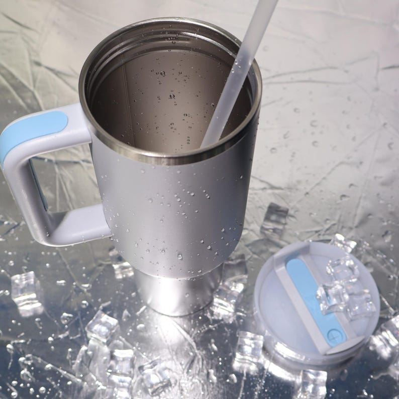 May include: A silver insulated travel mug with a light blue handle and lid. The mug has a stainless steel rim and a clear straw. The lid is light blue and white. The mug is sitting on a reflective surface with ice cubes.