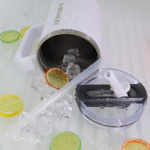 May include: A white Stanley insulated mug with a handle, filled with ice cubes. A clear straw and lid are next to the mug. Slices of lemon and orange are scattered around the mug, suggesting a refreshing drink.