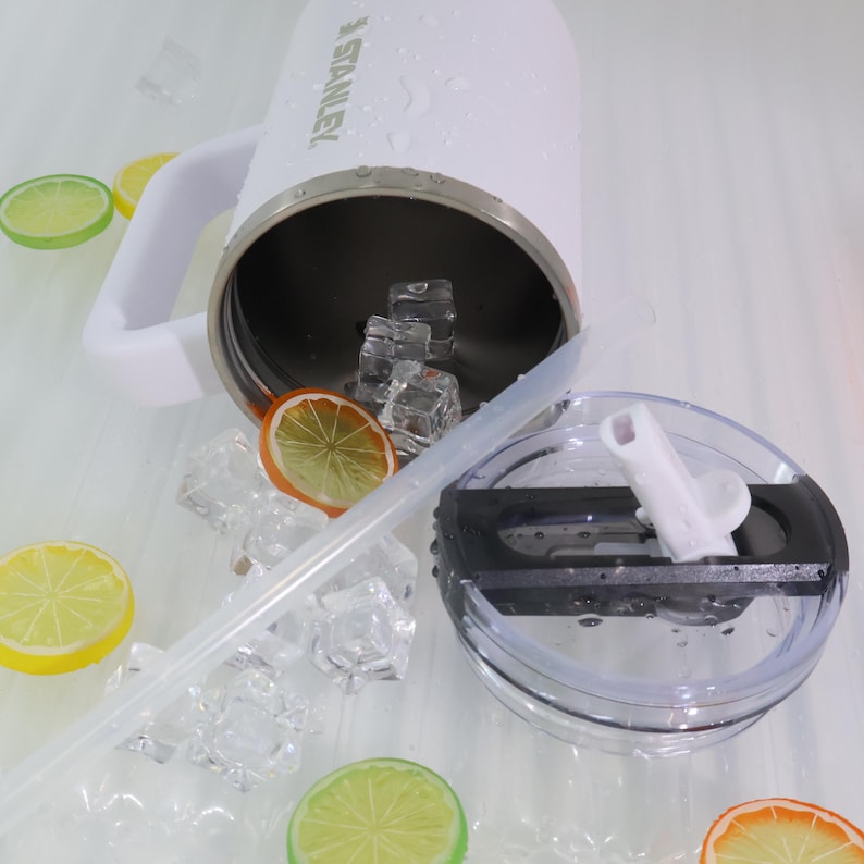 May include: A white Stanley insulated mug with a handle, filled with ice cubes. A clear straw and lid are next to the mug. Slices of lemon and orange are scattered around the mug, suggesting a refreshing drink.