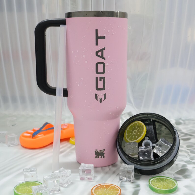 May include: A pink insulated tumbler with a black handle and the word "GOAT" printed on the side. The tumbler has a straw and is next to a lid with ice and lemon slices. A small orange flip-flop is in the background.