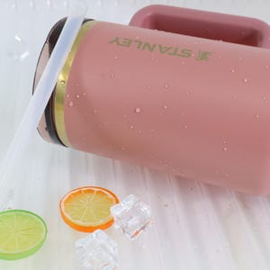 May include: A pink Stanley tumbler with a handle, a gold band, and the brand name in green. The tumbler is wet with water droplets. Slices of orange and lime, and ice cubes are next to the tumbler.