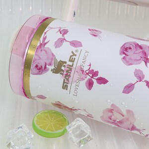 Pode incluir: Caneca Stanley Loveshack Fancy branca com estampa floral de rosas cor-de-rosa. A caneca tem uma tampa rosa e detalhes dourados. Uma fatia de lim&atilde;o e cubos de gelo est&atilde;o ao lado da caneca.