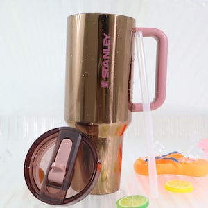 Puede incluir: Un vaso Stanley dorado con un asa rosa y el nombre de la marca impreso en rosa. El vaso incluye una tapa rosa a juego y una pajita transparente. El vaso est&aacute; dise&ntilde;ado para bebidas fr&iacute;as.