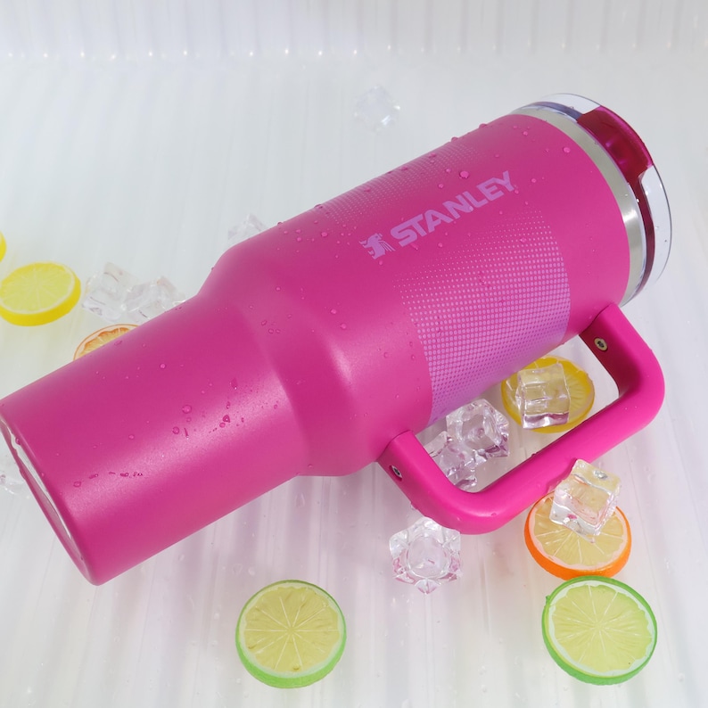Puede incluir: Un vaso Stanley rosa con asa, la marca es visible. El vaso est&aacute; rodeado de cubitos de hielo y rodajas de lim&oacute;n. Tiene un borde de acero inoxidable y una tapa transparente.