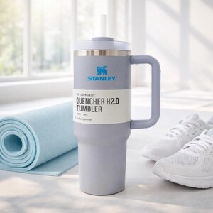 May include: A gray Stanley Quencher H2.0 tumbler with a handle and straw, next to a blue yoga mat and white sneakers. The tumbler has the Stanley logo and the text "QUENCHER H2.0 TUMBLER". It holds 40 oz (1.18L).