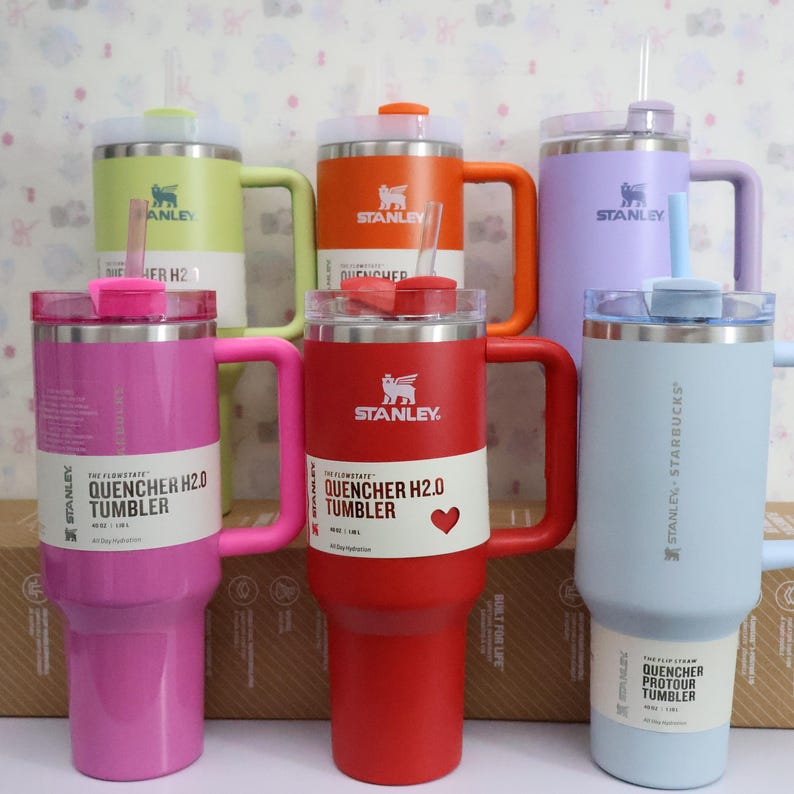 Puede incluir: Varios vasos Stanley Quencher H2.0 en colores como rosa, rojo, naranja, verde, morado y azul claro. Cada vaso tiene un asa, una pajita y el logotipo de Stanley. El texto "QUENCHER H2.0 TUMBLER" es visible en las etiquetas.