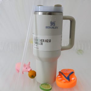 May include: A white Stanley Quencher H2.0 tumbler with a gray handle and lid. The tumbler has the Stanley logo and the text "QUENCHER H2.0 TUMBLER". A small pink pig, a small orange, a lime slice, and an orange flip-flop are in the foreground.