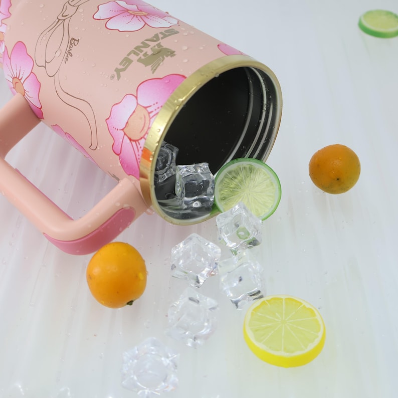 K&ouml;nnte beinhalten: Rosa Stanley-Reisebecher mit floralem Design und dem Wort "Barbie". Der Becher hat einen goldenen Rand und einen rosa Griff. Eisw&uuml;rfel, Orangenscheiben und Zitronenscheiben sind um den Becher verstreut.