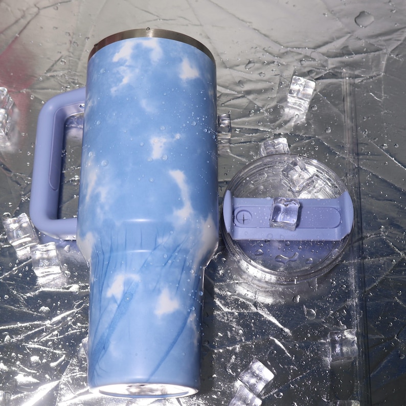 K&ouml;nnte beinhalten: Ein gro&szlig;er, isolierter Becher mit einem hellblauen und wei&szlig;en Wolkenmuster. Der Becher hat einen Griff und einen passenden hellblauen Deckel mit einer transparenten Kunststoffoberseite. Eisw&uuml;rfel und Wassertropfen umgeben den Becher.