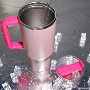 May include: A pink insulated travel mug with a bright pink handle and lid. The stainless steel mug has a silver rim and a clear lid with a pink handle. A clear straw is inside the mug. The mug is sitting on a reflective surface with ice cubes.