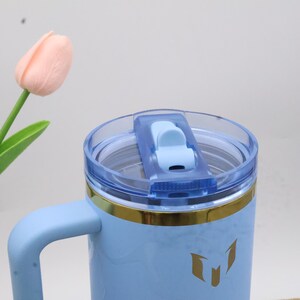 Pu&ograve; includere: Una tazza da viaggio azzurra con manico e coperchio trasparente. La tazza ha una fascia dorata e un'emblema dorato. Un tulipano rosa chiaro &egrave; sullo sfondo.