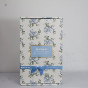 May include: A rectangular box with a floral and bow pattern in light blue, white, and green. The box features the text "STANLEY" and "LOVESHACKFANCY" in a blue rectangular label. A blue ribbon is tied around the box.