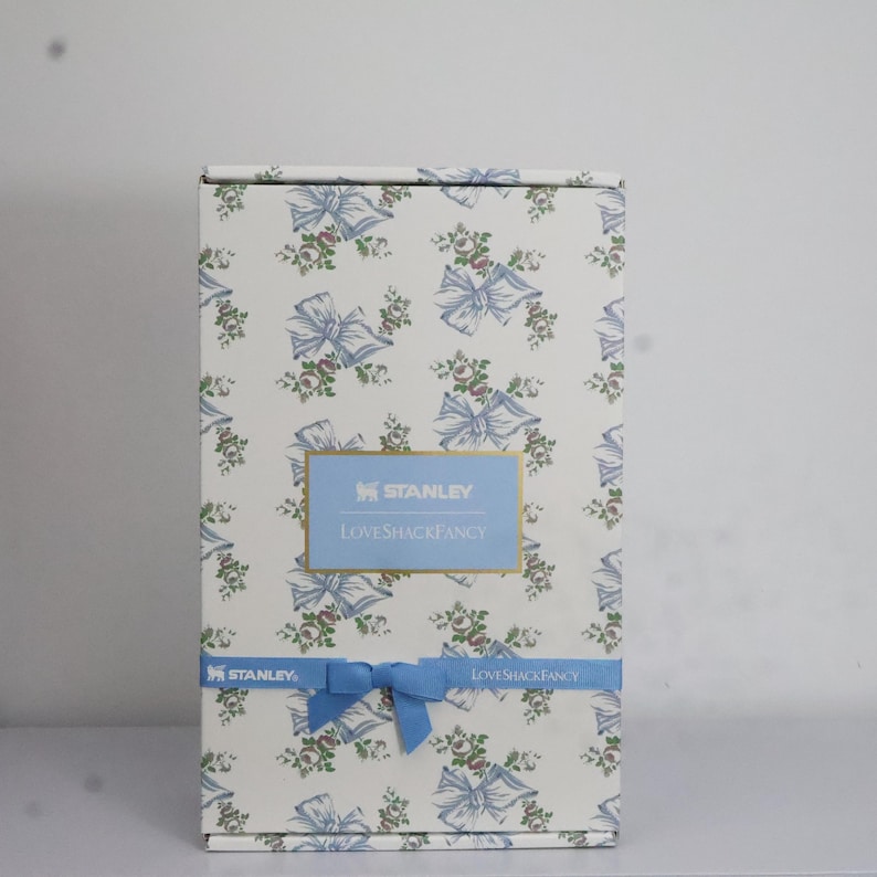 May include: A rectangular box with a floral and bow pattern in light blue, white, and green. The box features the text "STANLEY" and "LOVESHACKFANCY" in a blue rectangular label. A blue ribbon is tied around the box.
