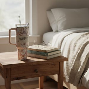 K&ouml;nnte beinhalten: Ein marmoriertes Trinkgef&auml;&szlig; mit Griff und Strohhalm steht auf einem Nachttisch aus Holz. Auf dem Becher steht der Text "QUENCHER H2.0 TUMBLER" und ein Logo. Daneben liegen B&uuml;cher und runde Brillen. Die Szene spielt in einem Schlafzimmer.