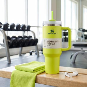 Op de afbeelding: Een limoengroene Stanley Quencher H2.0 tumbler met een witte deksel en handvat, en een bijpassend rietje. De tumbler heeft het Stanley-logo en productinformatie op een wit label. Een opgevouwen groene handdoek en witte oortelefoons liggen op een houten bank.
