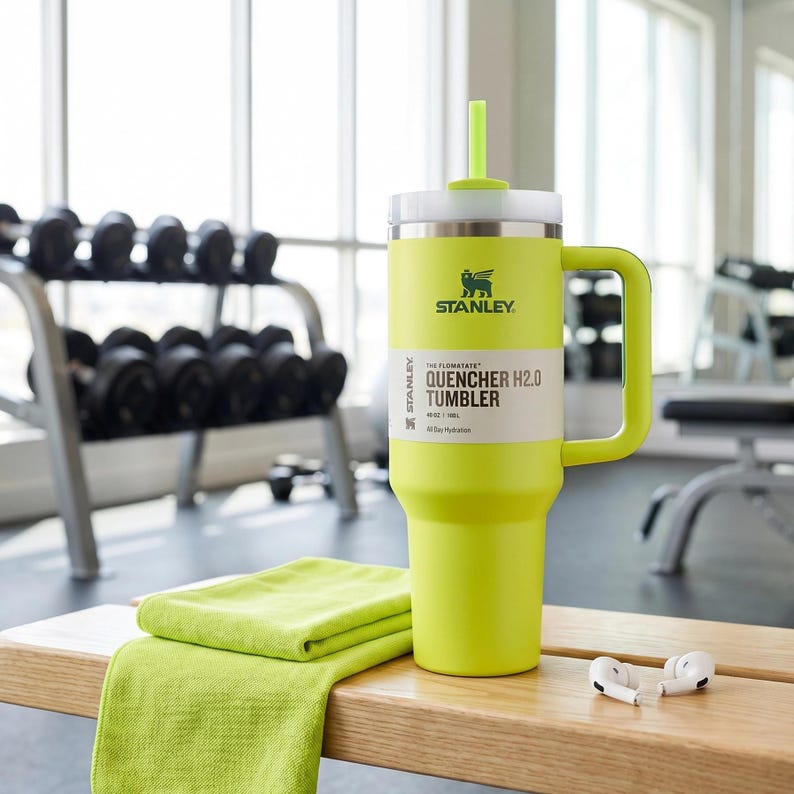 Op de afbeelding: Een limoengroene Stanley Quencher H2.0 tumbler met een witte deksel en handvat, en een bijpassend rietje. De tumbler heeft het Stanley-logo en productinformatie op een wit label. Een opgevouwen groene handdoek en witte oortelefoons liggen op een houten bank.
