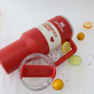 Puede incluir: Un vaso Stanley Quencher H2.0 rojo con un asa y tapa rojas a juego. El vaso tiene el logotipo de Stanley y la informaci&oacute;n del producto impresa. La tapa es transparente con un deslizador rojo. Limones decorativos y cubitos de hielo est&aacute;n esparcidos alrededor del vaso.