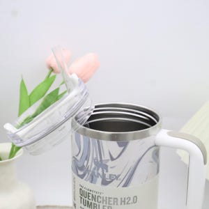 May include: A white and grey marble-effect Stanley Quencher H2.0 tumbler with a clear lid and straw. The tumbler has a white handle and a silver rim. The text "THE FLOWSTATE" and "QUENCHER H2.0 TUMBLER" are visible on the label.