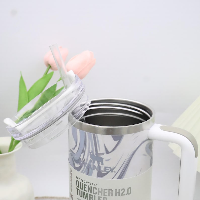 May include: A white and grey marble-effect Stanley Quencher H2.0 tumbler with a clear lid and straw. The tumbler has a white handle and a silver rim. The text "THE FLOWSTATE" and "QUENCHER H2.0 TUMBLER" are visible on the label.