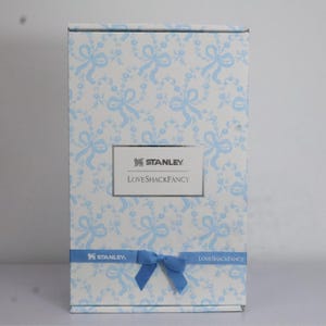 K&ouml;nnte beinhalten: Eine rechteckige Schachtel mit einem hellblauen und wei&szlig;en Blumenmuster und hellblauen Schleifen. Die Schachtel hat die Logos "STANLEY" und "LoveShackFancy". Ein hellblaues Band mit einer Schleife ist um die Schachtel gewickelt.
