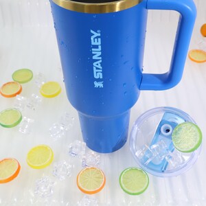 May include: A blue Stanley insulated tumbler with a handle, gold rim, and the brand name printed on the side. The tumbler is surrounded by ice cubes and decorative citrus fruit slices. A clear lid with ice and a citrus slice is also present.