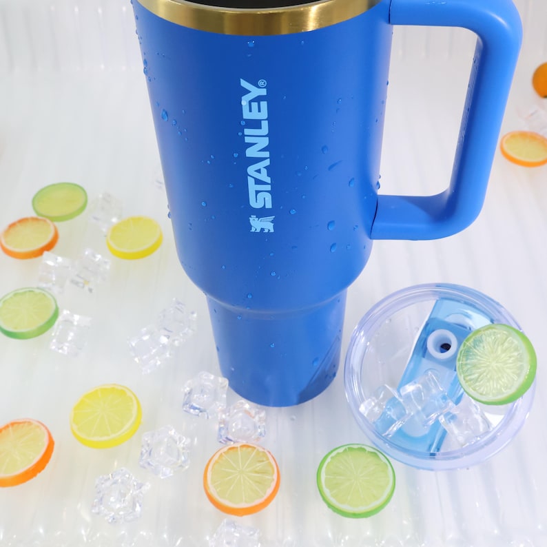 May include: A blue Stanley insulated tumbler with a handle, gold rim, and the brand name printed on the side. The tumbler is surrounded by ice cubes and decorative citrus fruit slices. A clear lid with ice and a citrus slice is also present.