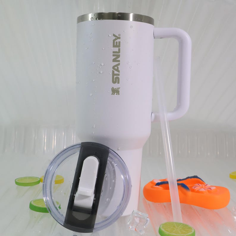 May include: A white Stanley insulated travel mug with a handle and a clear lid. The mug has the Stanley logo and a straw. The lid is off to the side. There is an orange flip-flop and lime slices in the background.