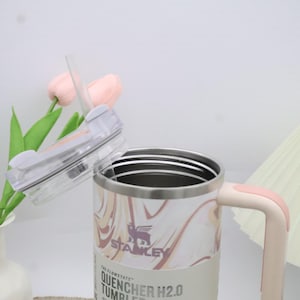 May include: A stainless steel tumbler with a marble pattern in shades of pink and cream. It has a pink handle and a clear lid with a straw. The tumbler has the text "STANLEY" and "QUENCHER H2.0 TUMBLER" on the side.