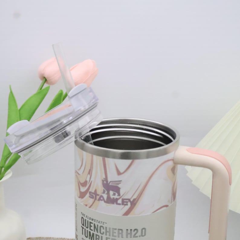 May include: A stainless steel tumbler with a marble pattern in shades of pink and cream. It has a pink handle and a clear lid with a straw. The tumbler has the text "STANLEY" and "QUENCHER H2.0 TUMBLER" on the side.