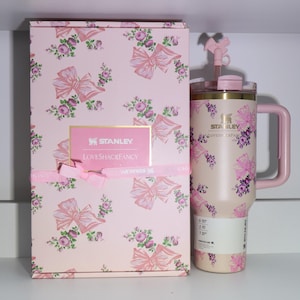 K&ouml;nnte beinhalten: Eine rosa Stanley LoveShackFancy Reisemug und eine passende Geschenkbox. Die Tasse und die Box weisen ein Blumen- und Schleifenmuster auf. Die Tasse hat einen Griff und einen rosa Deckel mit einem schleifenf&ouml;rmigen Akzent. Die Box hat ein rosa Band und ein goldgerahmtes Etikett.