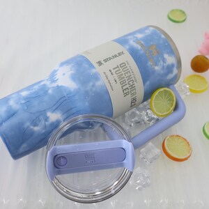 K&ouml;nnte beinhalten: Ein hellblau-wei&szlig;es Stanley Quencher H2.0 Tumbler mit passendem Griff und Deckel. Der Becher hat ein Fassungsverm&ouml;gen von 1,18 L. Der Deckel ist transparent mit einem hellblauen Schieber. Der Becher ist von Eisw&uuml;rfeln und dekorativen Fruchtscheiben umgeben.
