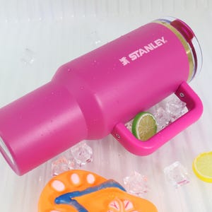 May include: A bright pink Stanley tumbler with a handle, the brand name in white. The tumbler is surrounded by ice cubes and a lime slice. The tumbler is designed to keep drinks cold.