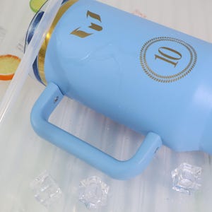 May include: A light blue insulated tumbler with a handle, gold accents, and a straw. The tumbler has a gold emblem and the number 10 in a gold circle. Ice cubes are visible near the tumbler.