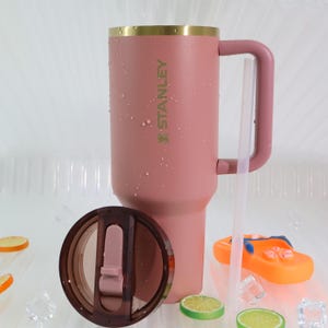 May include: A dusty rose Stanley tumbler with a handle, gold rim, and the brand name printed on the side. The lid is off, revealing the interior. A clear straw is next to the tumbler. The tumbler is sitting on a white surface.