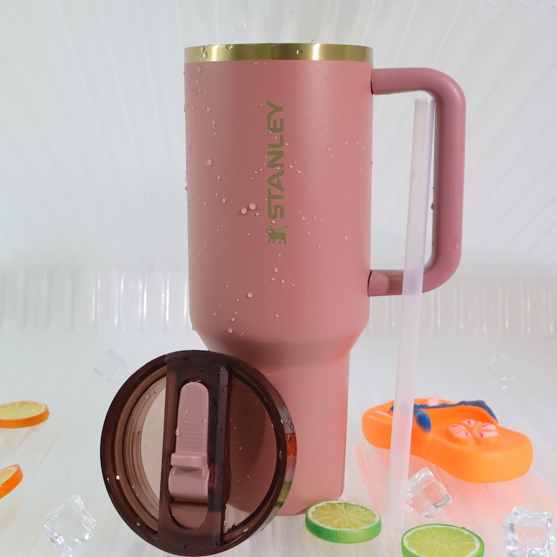 May include: A dusty rose Stanley tumbler with a handle, gold rim, and the brand name printed on the side. The lid is off, revealing the interior. A clear straw is next to the tumbler. The tumbler is sitting on a white surface.