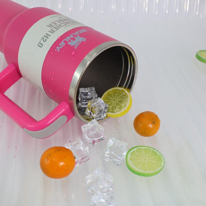 Pu&ograve; includere: Un bicchiere rosa Stanley Quencher H2.0 con manico, riempito con cubetti di ghiaccio, una fetta di limone, una fetta di lime e due arance. Il bicchiere ha una fascia bianca con il nome e il logo del marchio.