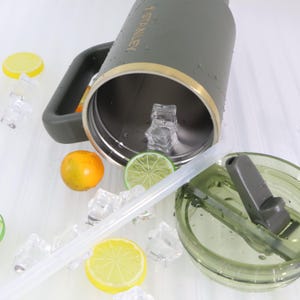 Puede incluir: Una botella de agua Stanley verde oliva con asa, ribete dorado y pajita transparente. La botella est&aacute; abierta, revelando el interior, y est&aacute; rodeada de cubitos de hielo, rodajas de lim&oacute;n y lima, y una naranja.