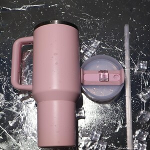 K&ouml;nnte beinhalten: Ein rosa isolierter Becher mit Griff, Deckel und Strohhalm. Der Becher ist zylindrisch mit einem Griff an der Seite. Der Deckel ist hellrosa mit einem transparenten Kunststoffschieber. Ein klarer Strohhalm liegt neben dem Deckel. Eisw&uuml;rfel sind verstreut.